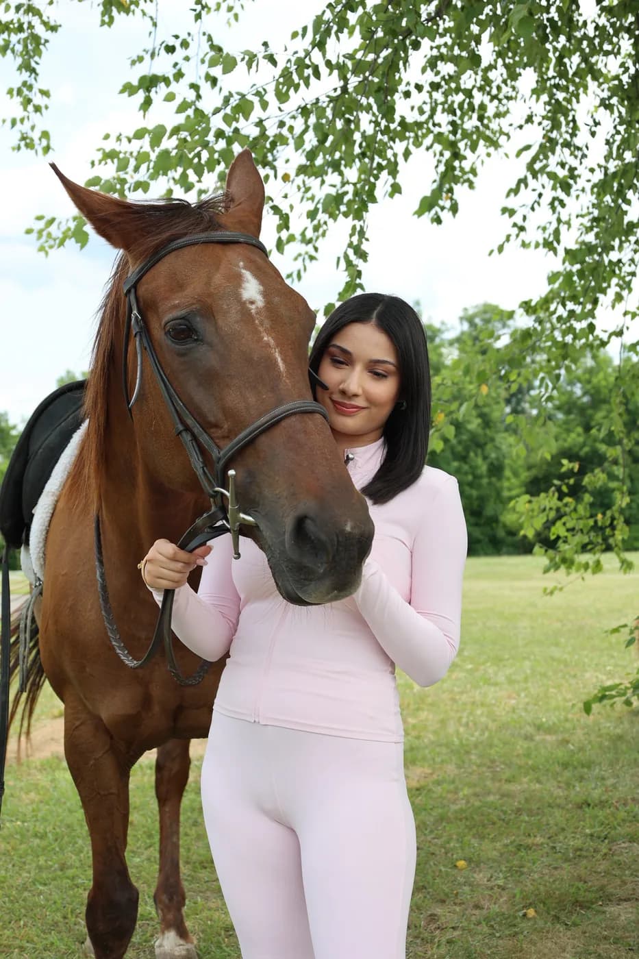 Guest in soft pink riding wear standing with a chestnut horse under birch trees at a Mind & Body Retreats event