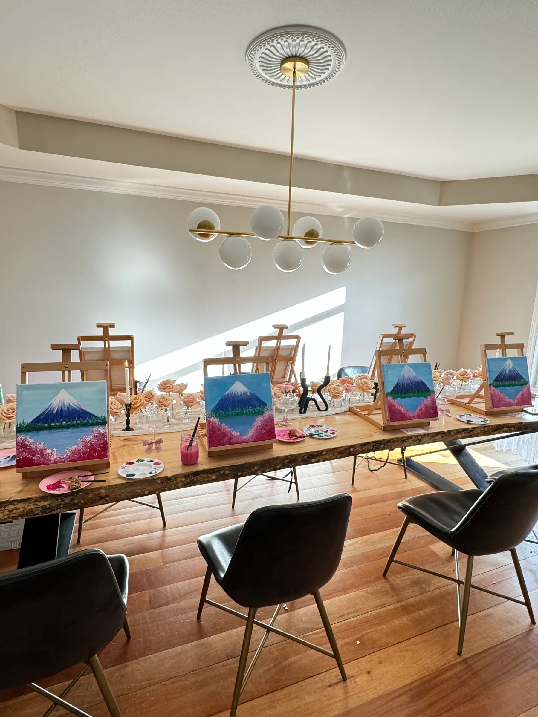 Easels holding painted mountain canvases lined down a long table with palettes and blush florals under a brass chandelier at a Mind & Body Retreats art workshop